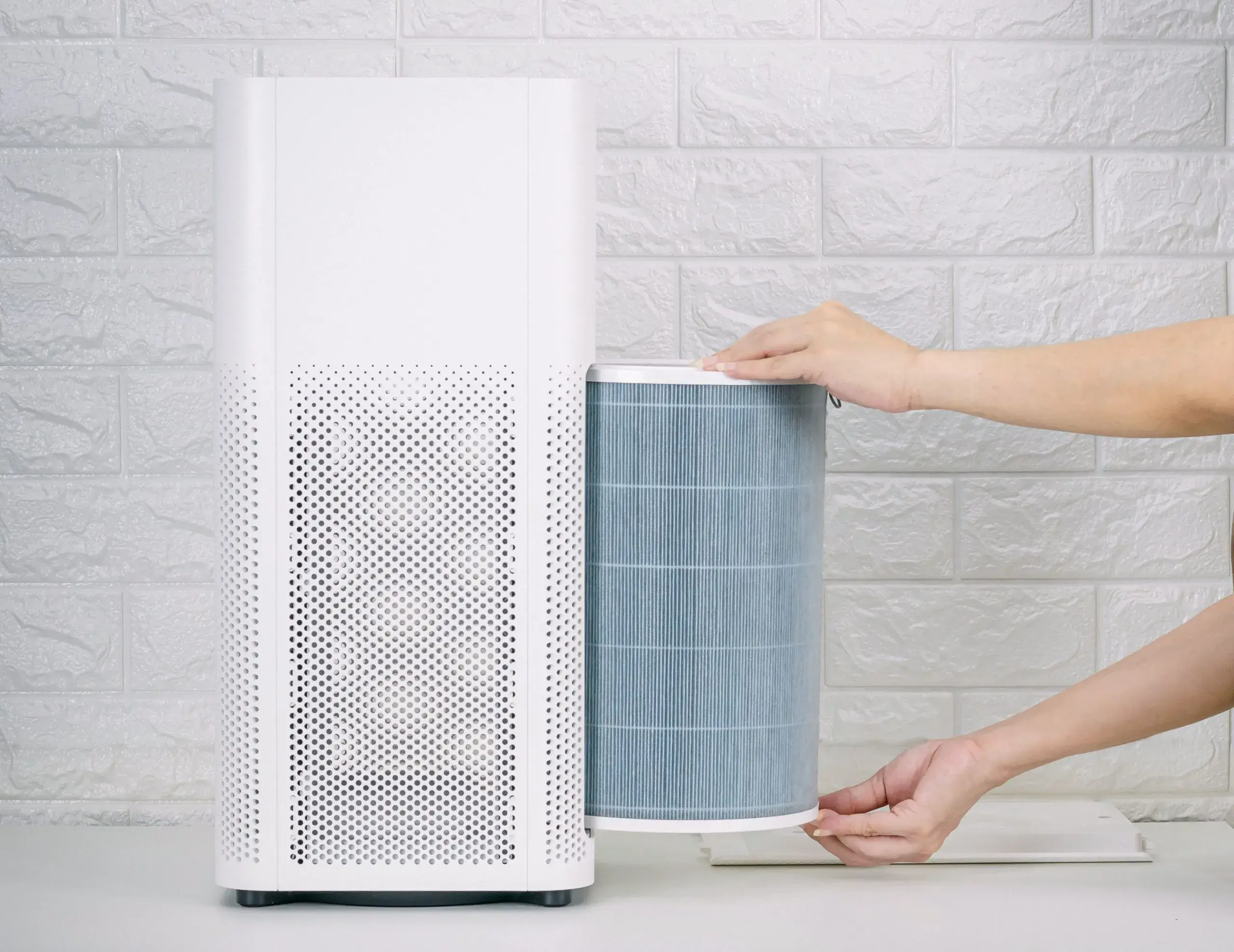 Person replacing a cylindrical air filter in a white air purifier against a white brick wall background, highlighting the importance of maintaining Indoor Air Quality in Raleigh, NC.