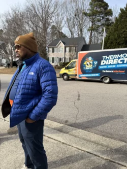 A man in a blue jacket and brown beanie stands on a suburban street near a parked Thermo Direct service van.